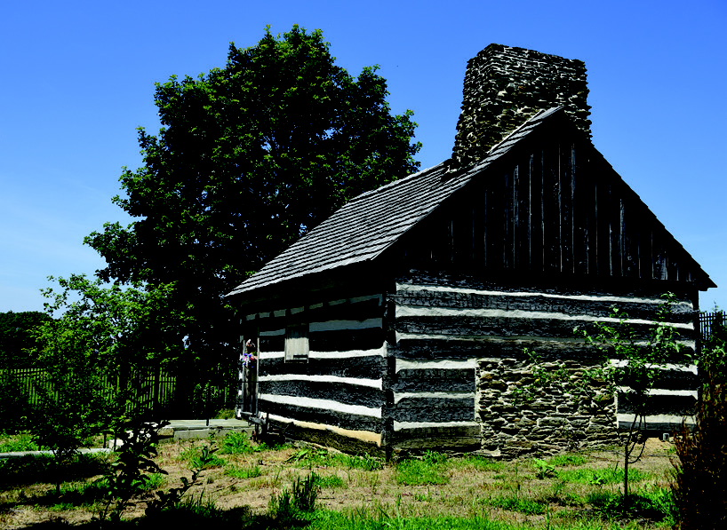 Rear exterior of the Neill Log House highlighting the original stone chimney, hand-hewn logs, and protective black aluminum picket fence.