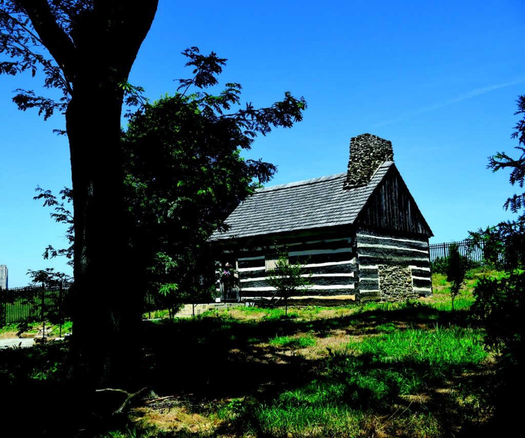 Wide shot of the historic Neill Log House nestled in the greenery of Schenley Park under a clear blue sky.