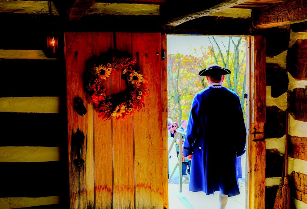 A historical reenactor in a blue colonial coat and tricorn hat walking through the front door of the Neill Log House, adorned with a floral wreath.