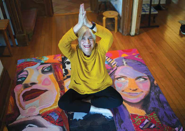 Artist Lauren Stern smiling and sitting cross-legged in a yoga pose on the floor between two large, colorful canvas paintings of women's faces.