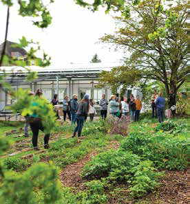 A large group of people gathered for an outdoor community educational workshop in a garden outside a modern greenhouse facility.