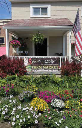 The storefront porch of The Corner Market featuring a large banner, hanging ferns, and vibrant flower beds out front.