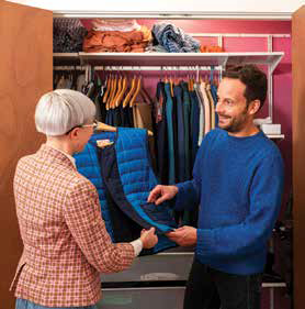 A professional wardrobe consultant helping a male client select a blue puffer vest during a home closet organization and styling service.