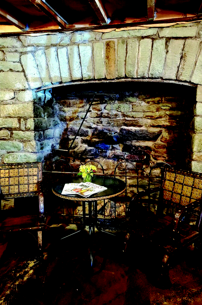 Interior of The Woods House showing a historic stone fireplace alcove with a small dining table, period-style chairs, and original timber beams.