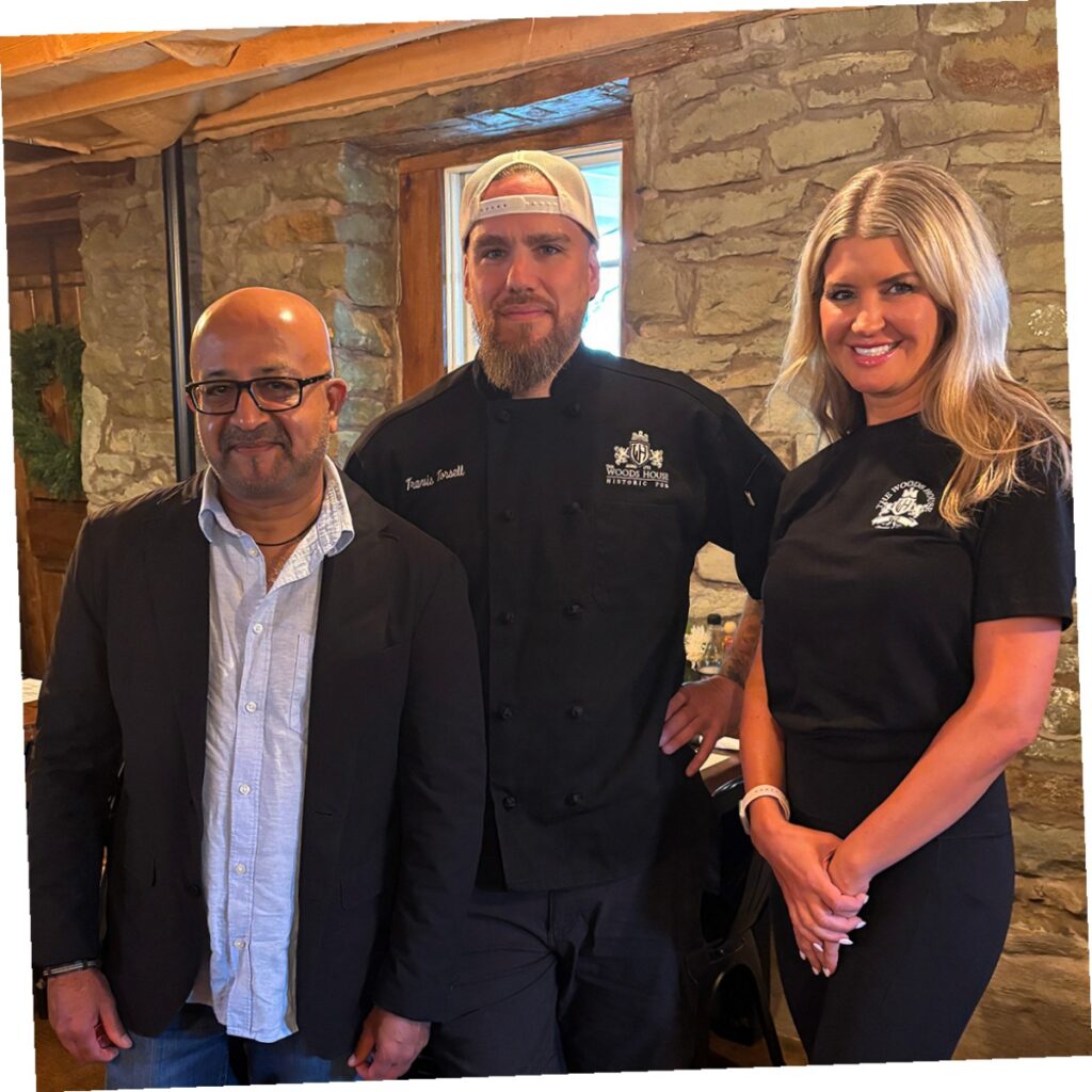 Owner Krish Pandya standing with Chef Travis Torsell and a staff member inside the historic stone interior of The Woods House Scottish pub.