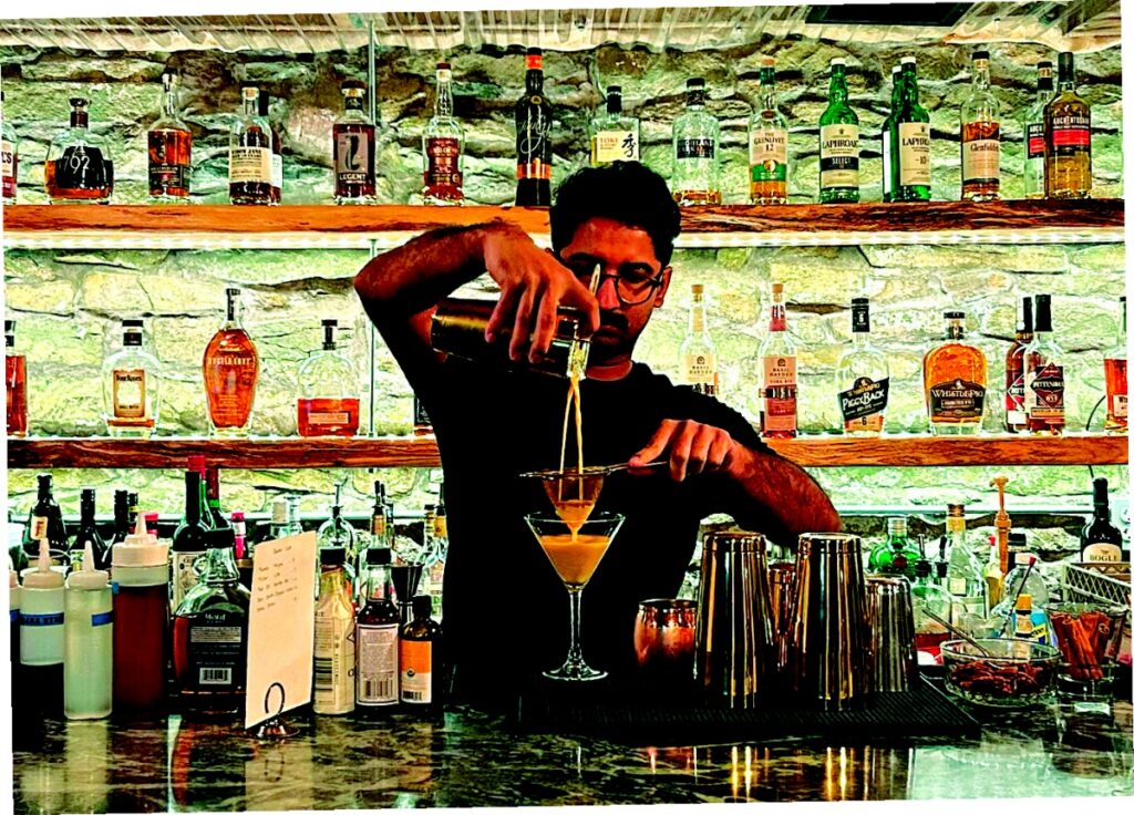 A bartender at The Woods House pouring a cocktail into a martini glass in front of a backlit bar stocked with local craft spirits.