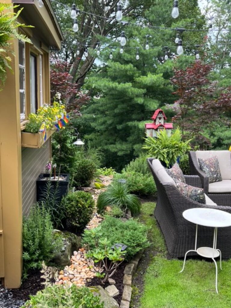 An outdoor patio seating area next to a custom potting shed and greenhouse with string lights in an urban garden.