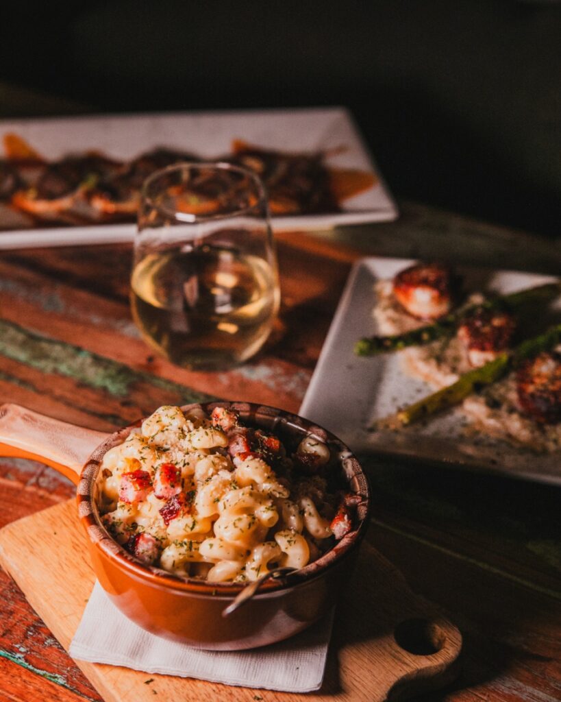 A bowl of gourmet mac and cheese with bacon served alongside white wine on a wooden table.