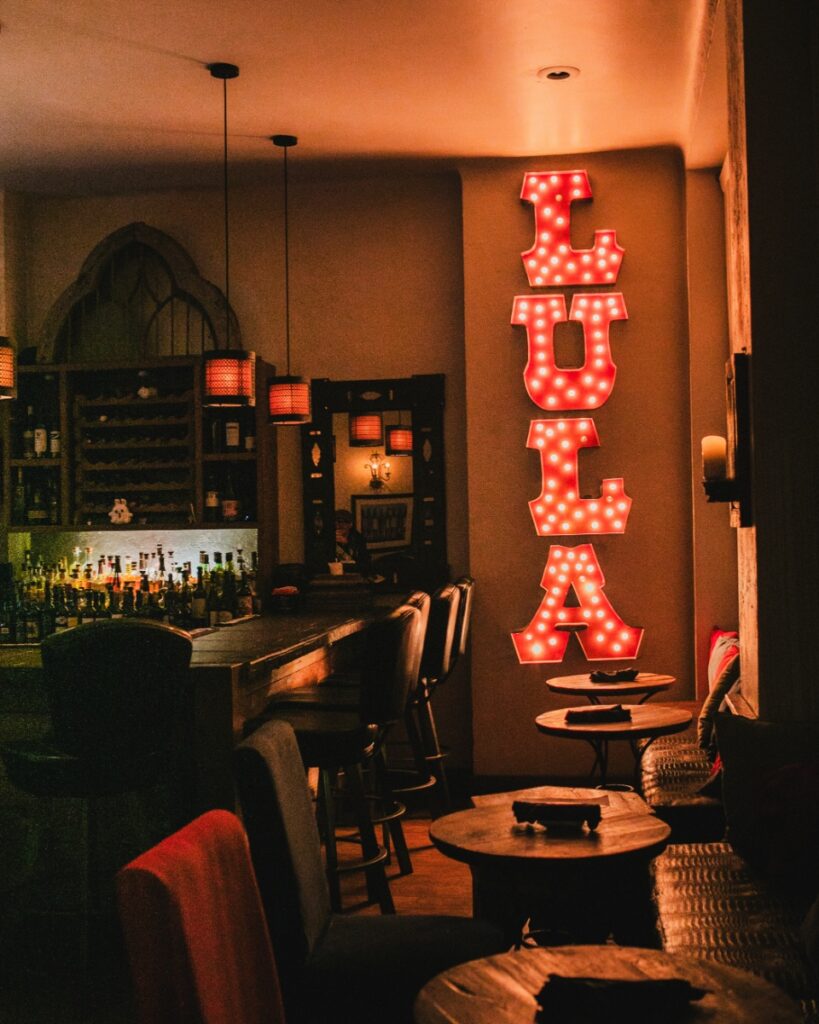 A cozy restaurant interior showing small tables, bar seating, and a large red neon Lula marquee sign.
