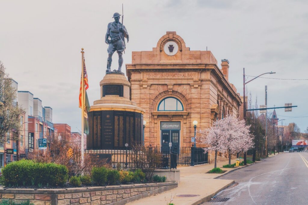 The historic Doughboy WWI monument at Doughboy Square in Lawrenceville, Pittsburgh, standing in front of the former Pennsylvania National Bank building with spring cherry blossoms.