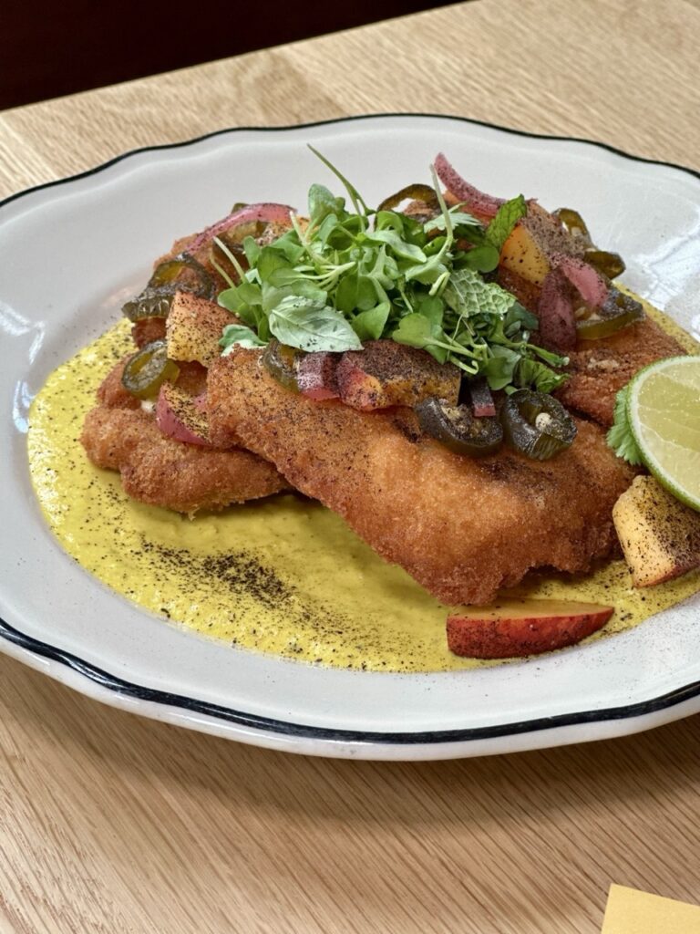 A white plate of chicken katsu with a yellow sauce below it and herbs on top.