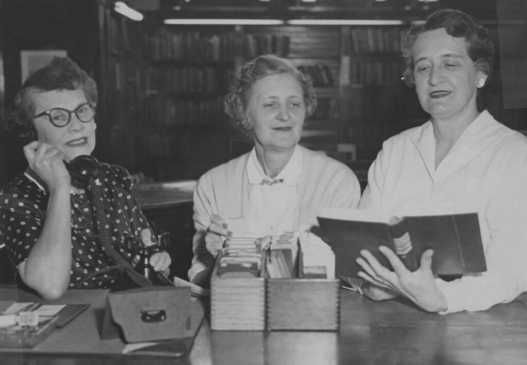 Three librarians stand at a desk of a library.