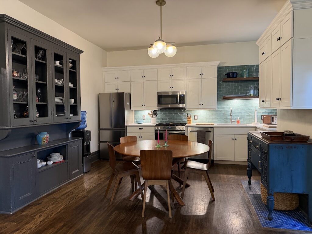 The inside of a kitchen and dining area with a dark blue cabinet to the left, a table in the middle, and a counter and cooking appliances behind the table.