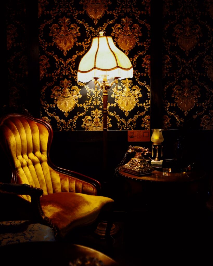Vintage yellow velvet armchair next to a fringed floor lamp and rotary phone against damask wallpaper.