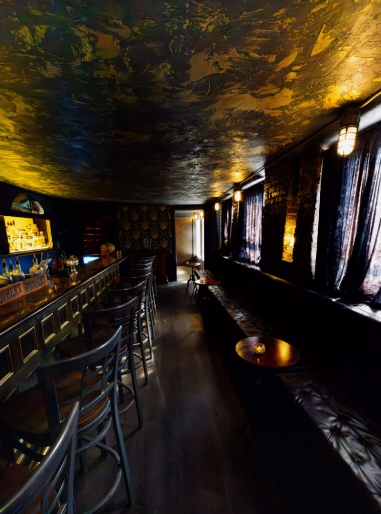 Dimly lit speakeasy lounge with dark leather banquettes, bar seating, and textured ceiling finish.