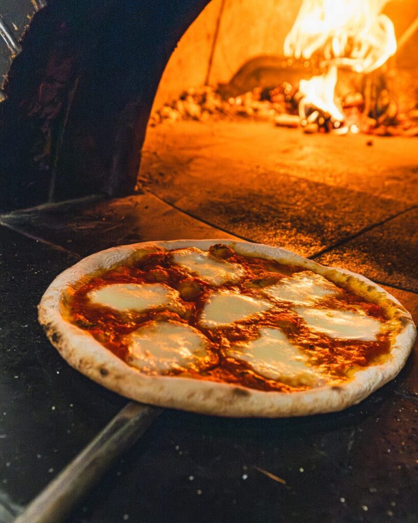 Freshly baked Margherita pizza on a peel coming out of a fiery wood-fired oven.