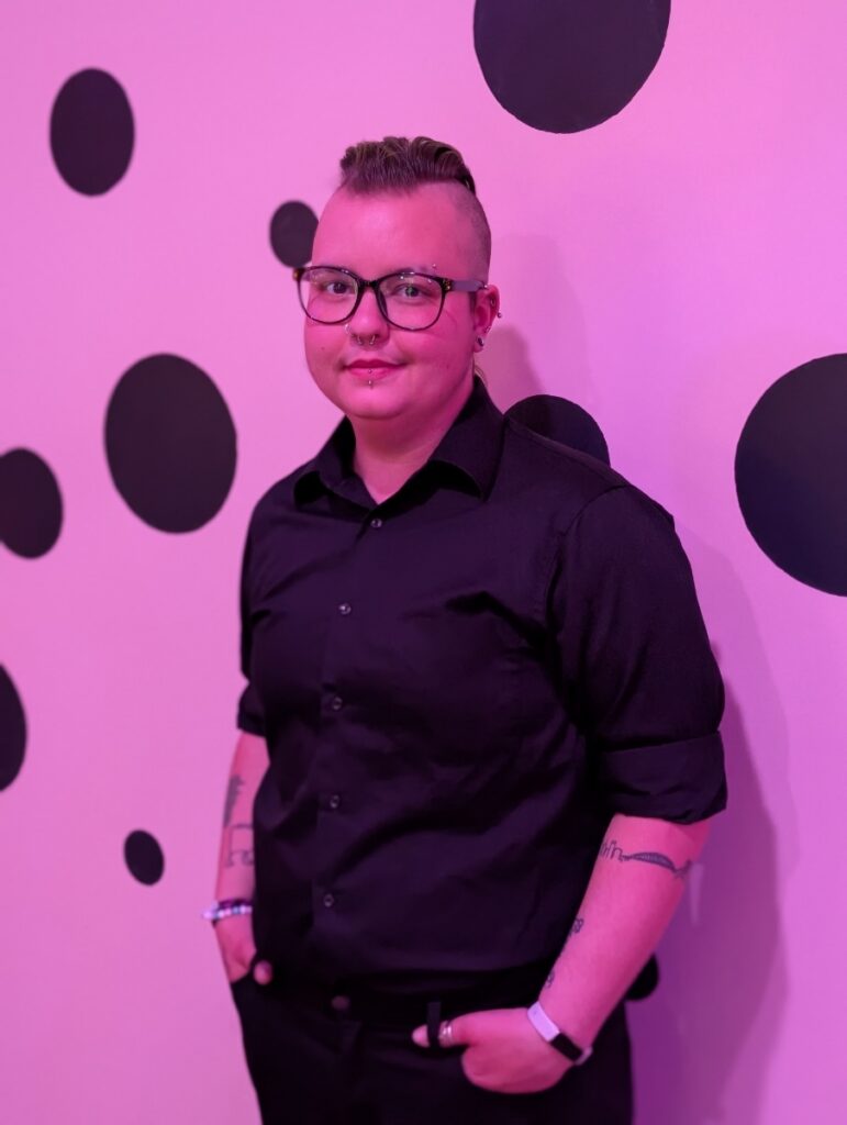 Portrait of Asmodeus Greer wearing glasses and a black button-up shirt in front of a pink polka-dot wall.