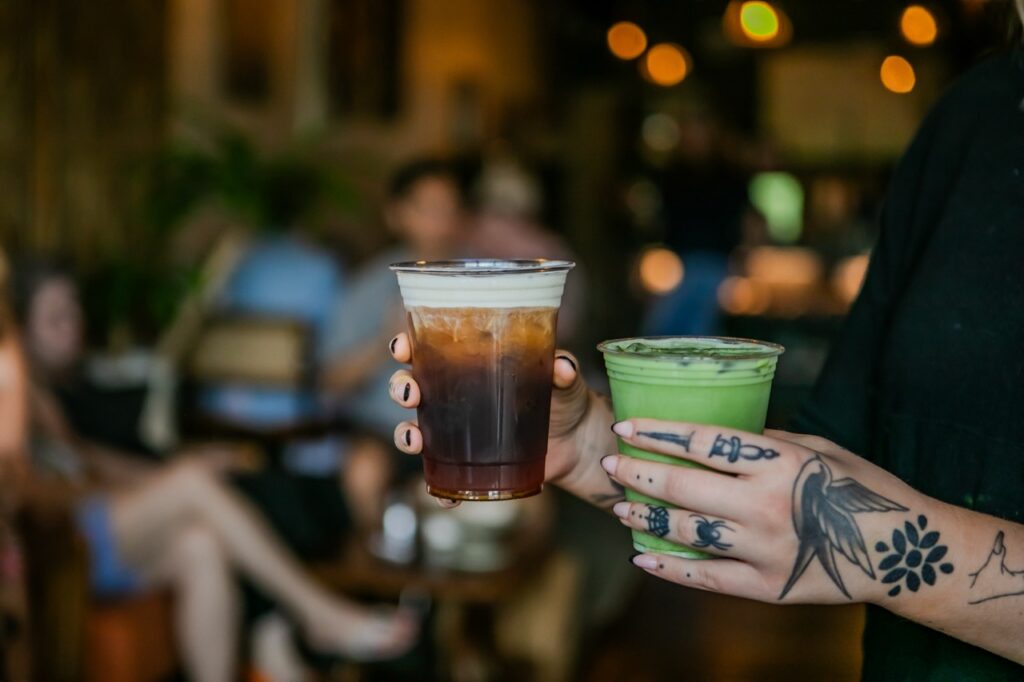 A person holds a latte and a matcha in their hands in a coffee shop.