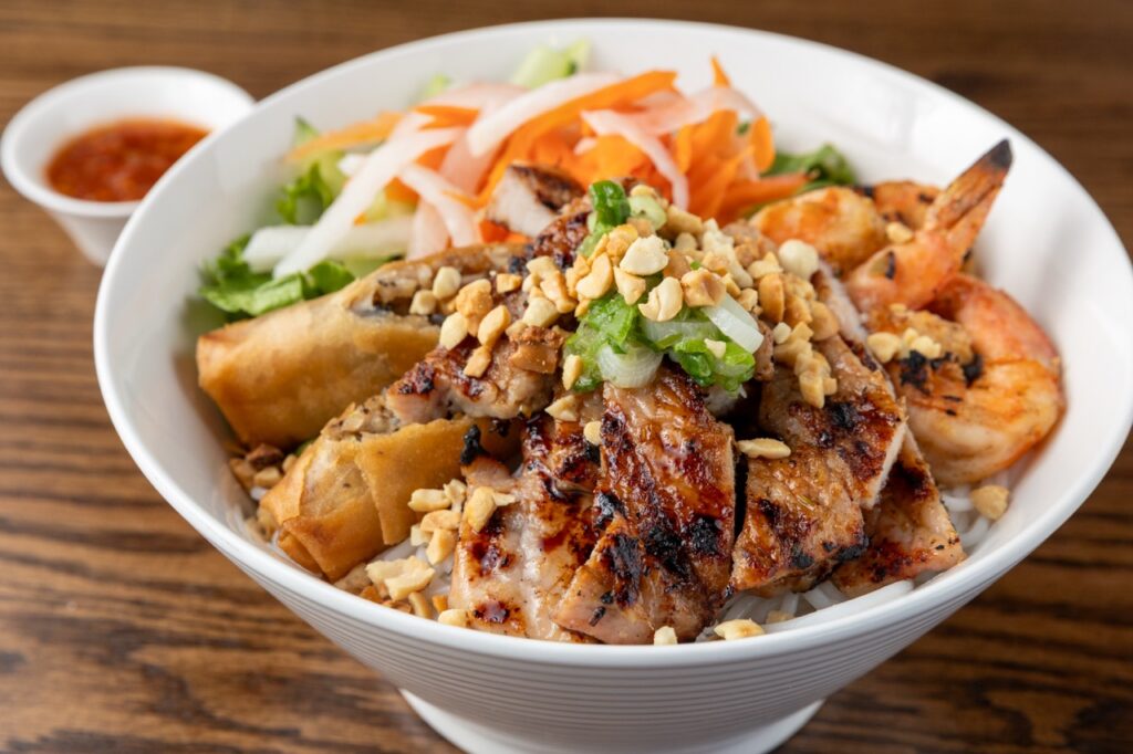 A bowl full of egg rolls, pork, vegetables, and shrimp with a small cup of red sauce beside it.