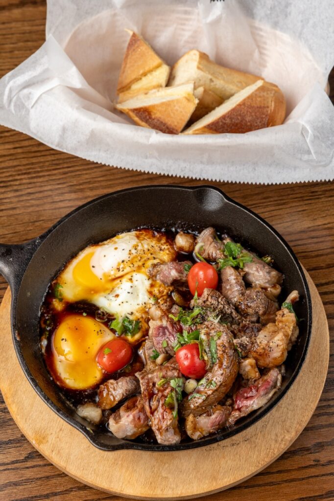 A skillet of steak and eggs below a basket of bread.