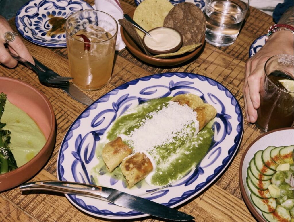 A blue and white plate with two long taquitos with sauce over top on a table full of other Oaxacan cuisine.
