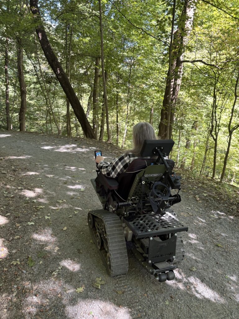 A woman uses a trackchair to move through a paved path in the woods.