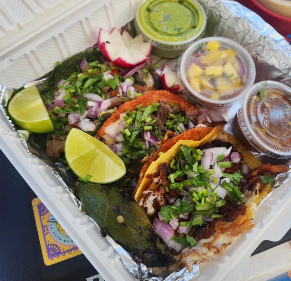 A container full of three tacos with limes nearby and three small containers.